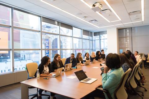 Multiple people seated at a long table with laptops open and turned on.