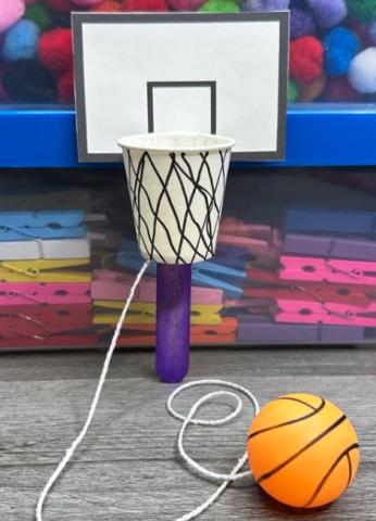 A small cup attached to a popsicle stick to make a hoop for a ping-pong ball tied to a string to make a toy. 